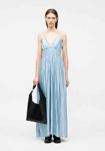 Femme portant une robe maxi plissée bleu clair à fines bretelles, tenant un grand sac à bandoulière noir et chaussée de chaussures noires.