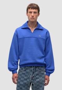 Felpa blu con colletto alto e una banda orizzontale cucita; realizzata in un tessuto morbido; abbinata a pantaloni in denim con motivo.