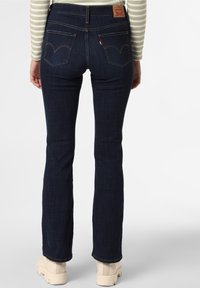 Jean évasé en denim bleu foncé avec une taille mi-haute, deux poches arrière et une étiquette en cuir marron. Associé à des bottes épaisses de couleur crème.