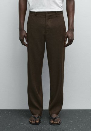 Man wearing dark brown loose pants, white shirt tucked in, and black sandals, standing against plain light gray wall.