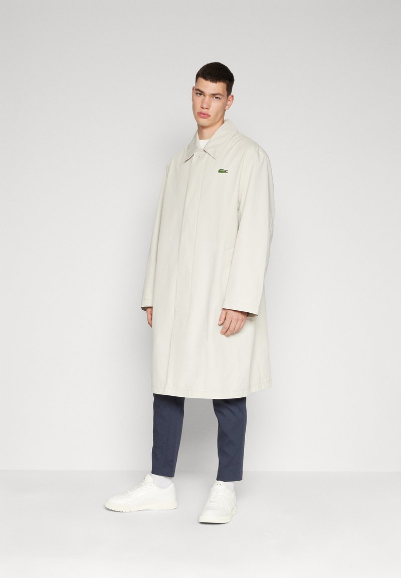 Impermeabile beige chiaro con taglio dritto, colletto chiuso e logo verde. Indossato sopra pantaloni blu navy e sneakers bianche, con una texture liscia.