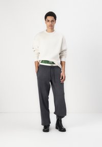 Pulover crem din tricot cu manșete cu nervuri, purtat cu pantaloni de trening largi în nuanța cărbunelui și cizme negre până la gleznă. Detaliu de tartan verde vizibil la talie.