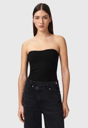 Jonge vrouw met steil bruin haar draagt een zwart ribgebreid topje zonder bandjes en hoge zwarte jeans tegen een effen grijze achtergrond.