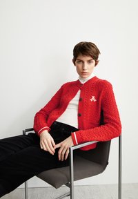 Rote Strickjacke mit strukturiertem Muster, rundem Ausschnitt, dunklen Knöpfen und weißen Akzenten. Getragen über einem weißen Oberteil und schwarzen Hosen.