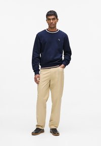 Sweat-shirt bleu marine à col rond avec bord-côtes rayé. Pantalon beige à coupe décontractée et chaussures noires.