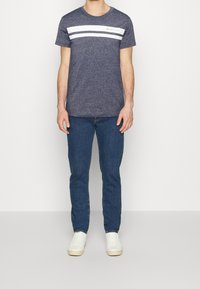 Mężczyzna w granatowej koszulce z białymi poziomymi paskami, niebieskich dżinsach i białych trampkach, stojący na tle czystej białej ściany.