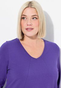 Lila Langarmshirt mit V-Ausschnitt, das eine glatte Textur und eine nahtdetail über die Vorderseite aufweist. Der Stoff wirkt weich und bequem.