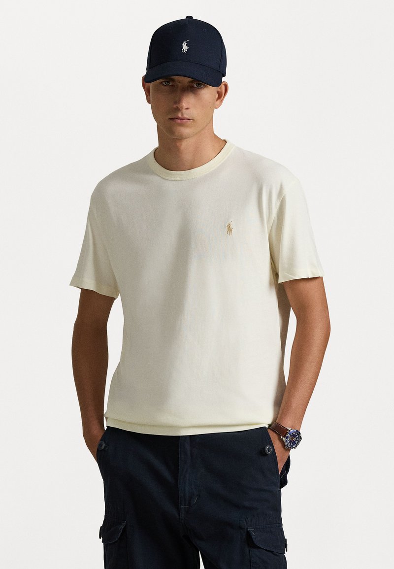 T-shirt de algodão amarelo claro com decote redondo e um pequeno logótipo bordado. Combinado com calças cargo escuras e um boné azul-marinho.