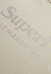 Sudadera beige con la palabra "Superdry" en rhinestones plateados, con "LE MARAIS PARIS" en una fuente simple debajo. Textura de tela suave.