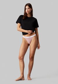 Zwarte korte mouwen t-shirt gecombineerd met een roze geribbelde thong met een witte tailleband met logo. Model staat blootsvoets met een neutrale uitdrukking.