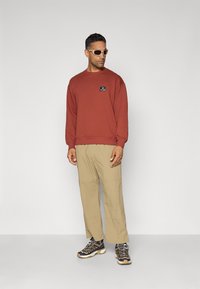 Felpa color ruggine con un piccolo logo sul petto, abbinata a pantaloni beige larghi e scarpe da trekking nere con accenti grigi.