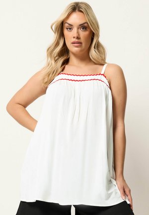 Lyshåret kvinde iført en løs, hvid, ærmeløs top med rød bølget kant ved halsudskæringen, stående med den ene hånd på hoften.