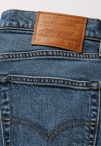 Denimjeans med en beige läderlapp. Lappen har präglad text och en logotyp, med gul sömnad längs sömmarna.