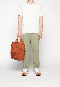 Béžové tričko s kapsou, zelené tepláky, bílé tenisky s oranžovými akcenty a oranžová plátěná taška s klopou a přezkou.