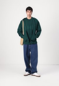 Grønn zip-up hettegenser lagd over en grønn T-skjorte, kombinert med løse blå jeans og hvite sneakers med beige detaljer. Beige skulderveske.