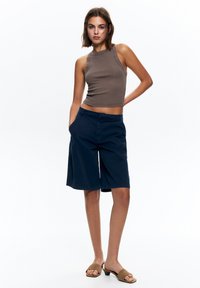 Hnedý rebrovaný crop top s vysokým výstrihom, skombinovaný s námorníckymi modrými šortkami s bočnými vreckami. Model má na sebe béžové nazúvacie sandále.