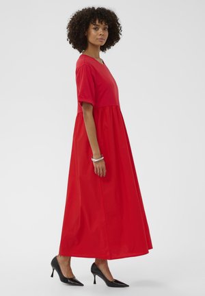 Kobieta w jaskrawo czerwonej sukni maxi z krótkimi rękawami i czarnymi szpiczastymi szpilkami, stojąca bokiem na jednolitym tle.