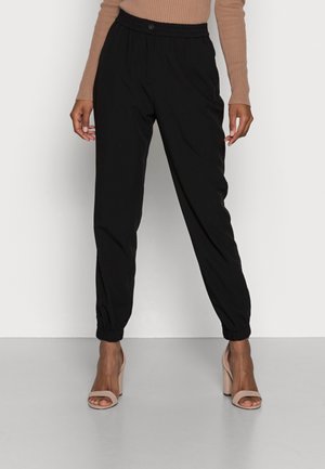 Mujer está de pie vistiendo pantalones negros ajustados con puños elásticos y sandalias beige de tacón alto, combinados con una camiseta beige de manga larga acanalada.