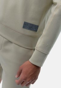 Beige sweatshirt with a neoprene effect, elastic fabric, and a small blue label. Features minimalist branding on the sleeve. Smooth texture.
