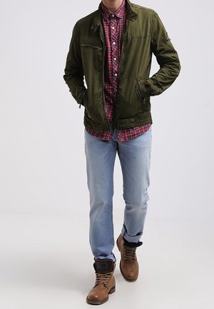 Olivgrön jacka med två sidofickor, lager på lager över en röd rutmönstrad skjorta, i kombination med ljusblåjeans och bruna snörstövlar.