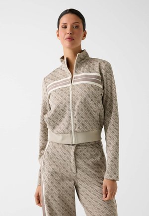 Kobieta stojąca, ubrana w beżową, wzorzystą kurtkę zapinaną na zamek oraz dopasowane spodnie z białymi paskami i nadrukiem logo na jednolitym białym tle.