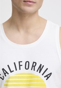 Top alb cu bretele, având cuvântul "CALIFORNIA" scris cu litere negre îndrăznețe și o grafică a soarelui galben cu dungi orizontale. Material din bumbac moale.
