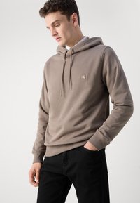 Sudadera con capucha marrón hecha de tela suave, con un pequeño parche de logo en el pecho, puños de canalé y un corte relajado. Combinada con pantalones negros.
