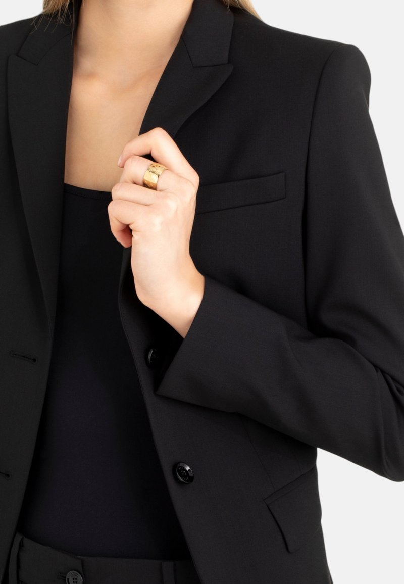 Blazer nero su misura con un unico bottone, rever a punta e una tasca frontale, abbinato a una maglia scura e con un anello d'oro sulla mano.