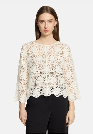 Jonge vrouw met steil bruin haar draagt een witte blouse van bloemenkant met driekwart mouwen en een zwarte broek, staand tegen een witte achtergrond.