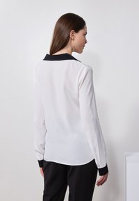 Bílá blůza s černým límcem a manžetami, dlouhé rukávy, hladký materiál, mírně průsvitná textura, přiléhavý střih a zakulacený lem.