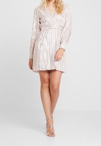 Robe décolletée en V plongeant avec des manches longues, en tissu argent plissé, taille cintrée et jupe évasée. Associée à des sandales à talons argentées.