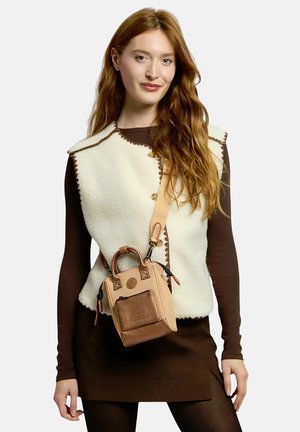 Frau mit langen roten Haaren trägt eine cremefarbene ärmellose Weste über einem braunen Kleid und trägt eine kleine braune Umhängetasche mit Fronttasche.