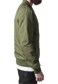 Veste bomber vert olive avec fermeture éclair, poignets et ceinture côtelés ; texture lisse et design épuré, vue de côté.