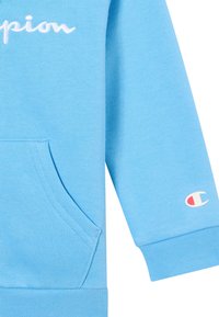Niebieska bluza z miękkiego materiału, z kieszenią kangurką, ściągaczami przy rękawach oraz haftowanym logo "Champion" na klatce piersiowej i rękawie.