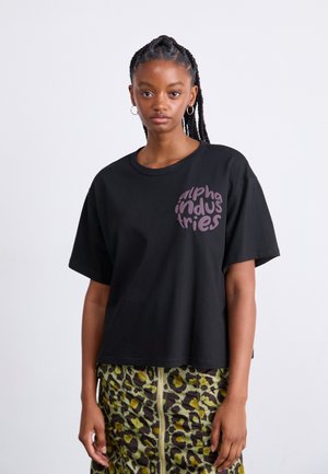 Camiseta de algodón negra con un corte holgado, que presenta un logo circular púrpura que dice "alpha industries" en el lado izquierdo del pecho.