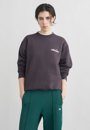 Mladá osoba s krátkymi tmavými vlasmi, oblečená v tmavosivej mikine ellesse a zelených teplákoch, stojaca s rukami v vreckách.