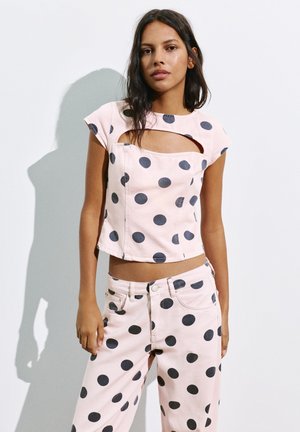 Donna che indossa un crop top rosa chiaro e pantaloni con grandi pois neri, in piedi su uno sfondo bianco.