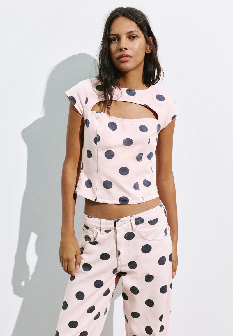 Femme portant un crop top rose clair et un pantalon à gros pois noirs, debout sur un fond blanc.