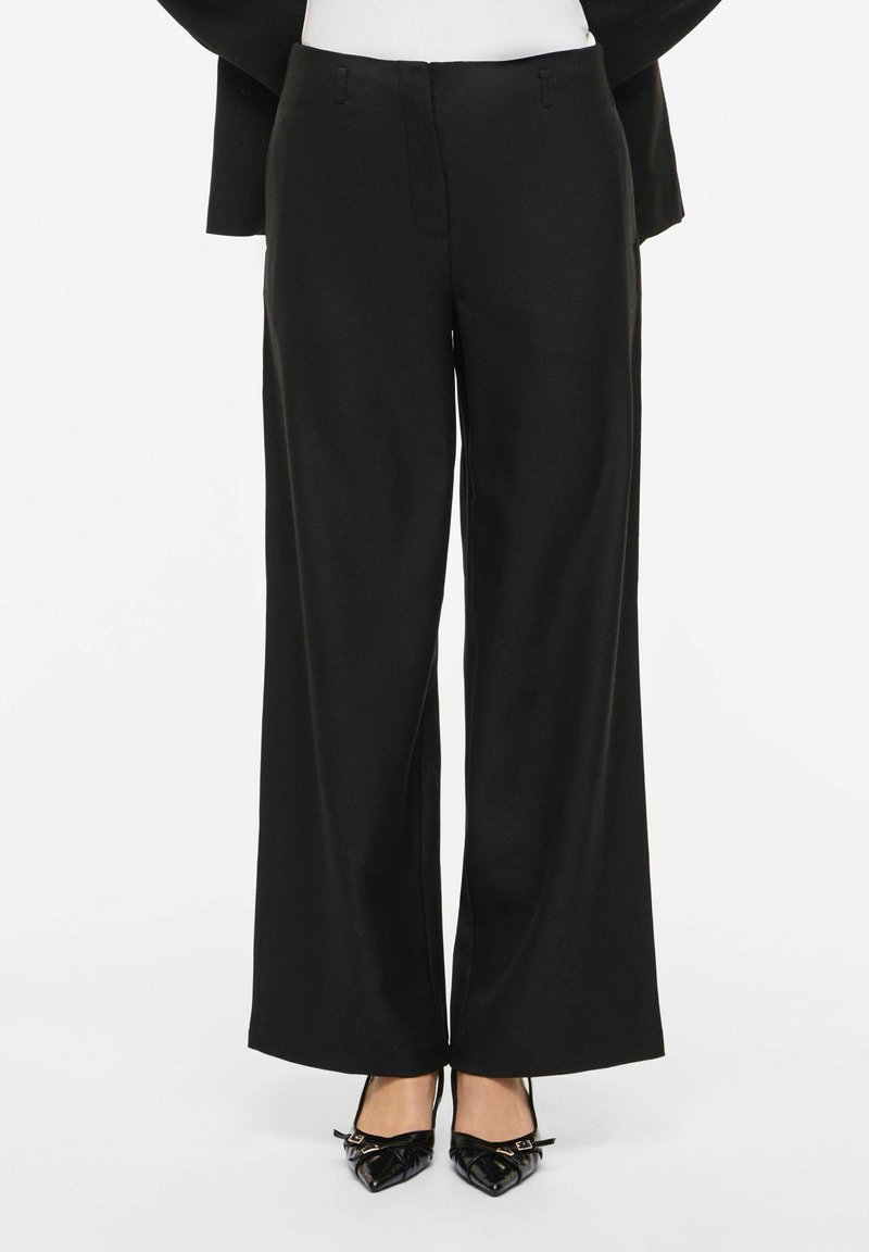 Črne hlače širokih nogavic iz gladkega blaga z urejenim sprednjim dizajnom in subtilnimi zankami za pas, kombinirane z ostrimi črnimi petami.
