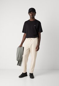 T-shirt noir à manches courtes avec logo, pantalon crème avec cordon de serrage, sweat-shirt gris à la main et baskets noires. Design épuré et couleurs neutres.
