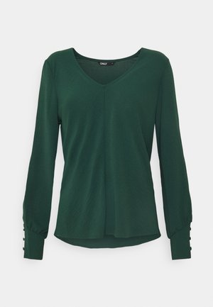 Blouse manches longues vert foncé avec col en V et poignets boutonnés, ourlet légèrement évasé, présentée sur fond uni.