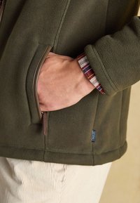 Hand in der Tasche einer olivgrünen Fleecejacke, die braune Lederbesätze und einen rot-weiß-blau karierten Hemdärmel über hellen Hosen zeigt.