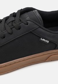 Schwarze Lederturnschuhe mit strukturiertem Obermaterial, schwarzen Schnürsenkeln und einer hellbraunen Gummisohle. Verfügt über dezente Markenkennzeichnung an der Seite.