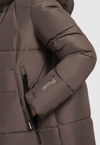 Puhasta jakna v rjavi tkanini s prešito zasnovo, ki ima zadrgo žep in tiskano logotipno podrobnost na rokavu.