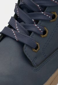 Zapato de cuero azul con cordones azul marino, ojales dorados y una superficie texturizada. La suela exterior es beige, añadiendo contraste al diseño.