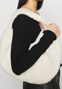 Cremefarbene Schultertasche aus Kunstlammfell mit abgerundeter Form und weicher Textur, ausgestattet mit einem breiten Riemen und minimalen Designdetails.