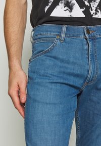 Jeans in denim blu con taglio dritto, caratterizzati da cuciture a contrasto, accessori in metallo e design a cinque tasche, tenuti su da una mano.