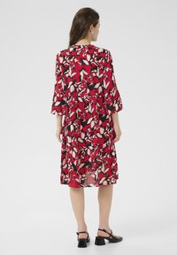 Robe florale rouge avec un design à étages, manches 3/4, avec des accents blancs et noirs. Modèle portant des sandales à talons noires.