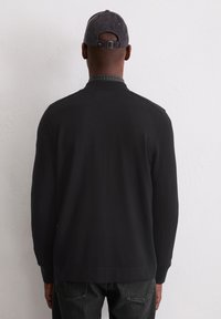 Schwarzer Pullover mit einem gerippten Kragen und langen Ärmeln, der eine glatte Textur aufweist. Wird über einem karierten Hemd getragen, Rückansicht.