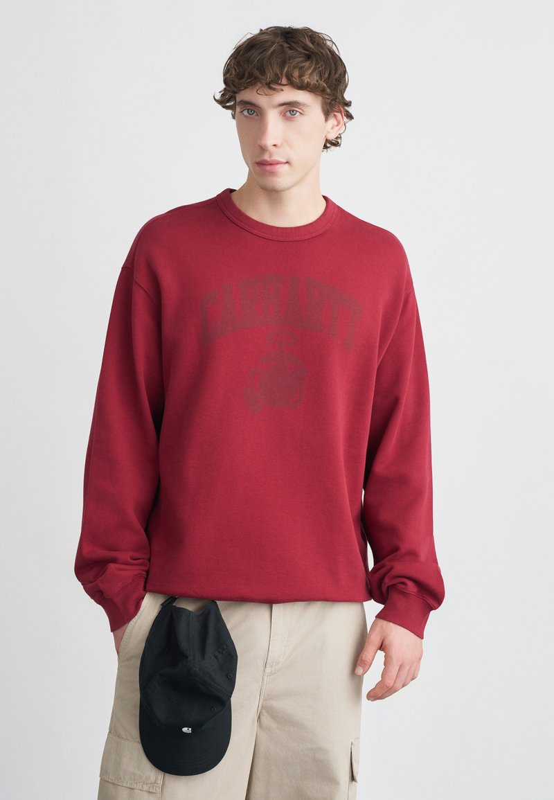 Sweatshirt rouge avec col rond côtelé, manches longues et un grand logo noir imprimé à l'avant. Porté avec un short cargo beige et une casquette noire.